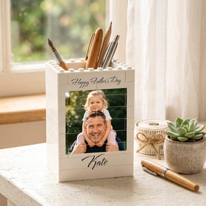 May include: A white rectangular pen holder constructed from interlocking plastic building blocks. The front features a printed photograph of a child sitting on an adult's shoulders, with the text Happy Father's Day above and the name Kate printed below.