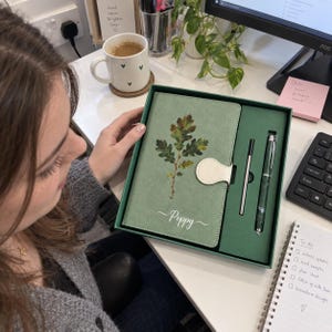 May include: A sage green notebook set in a matching box, featuring a printed oak leaf design and the name Poppy in white script. The set includes a notebook with a white magnetic closure, a slim metal pen, and a refill cartridge in a green tray.