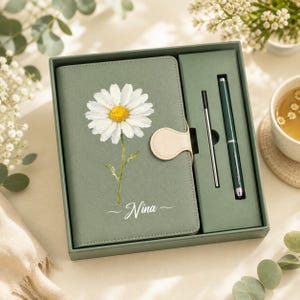 May include: A sage green notebook set featuring a white daisy illustration and the name Nina printed on the cover. The set includes a matching green pen and a thin metal refill, all housed within a rectangular green gift box with a beige closure.