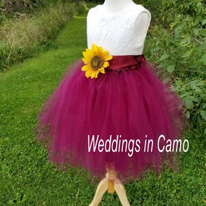 May include: A white lace bodice with a burgundy tulle skirt and a sunflower accent. The dress has a burgundy satin sash. The dress is on a wooden mannequin and is displayed against a green grassy background. The text "Weddings in Camo" is visible at the bottom of the image.