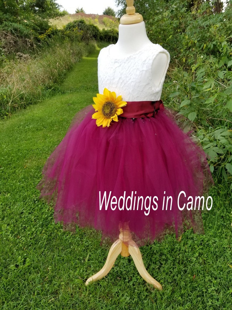 May include: A white lace bodice with a burgundy tulle skirt and a sunflower accent. The dress has a burgundy satin sash. The dress is on a wooden mannequin and is displayed against a green grassy background. The text "Weddings in Camo" is visible at the bottom of the image.
