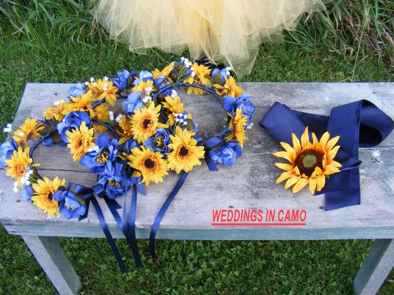 May include: Three sunflower and blue flower wreaths with navy blue ribbons, and a single sunflower with navy blue ribbon. The text "WEDDINGS IN CAMO" is visible on a wooden surface.