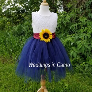 May include: A navy blue tulle flower girl dress with a white lace bodice and a burgundy sash. A yellow sunflower is attached to the sash. The dress is on a wooden mannequin and the text "Weddings in Camo" is visible at the bottom of the image.