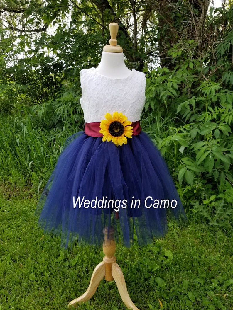 May include: A navy blue tulle flower girl dress with a white lace bodice and a burgundy sash. A yellow sunflower is attached to the sash. The dress is on a wooden mannequin and the text "Weddings in Camo" is visible at the bottom of the image.