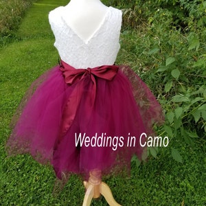 May include: A white lace bodice with a burgundy tulle skirt and a burgundy satin sash. The dress is on a mannequin and the background is green grass.