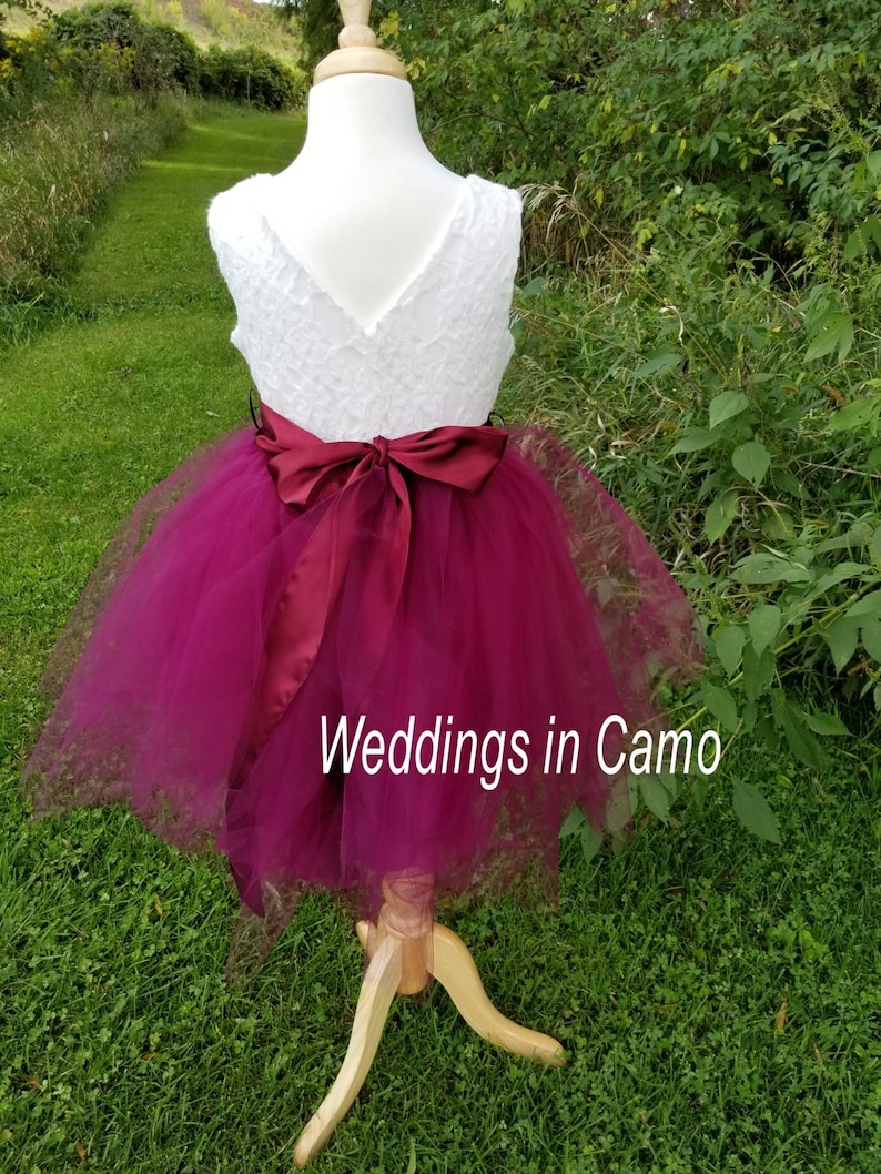 May include: A white lace bodice with a burgundy tulle skirt and a burgundy satin sash. The dress is on a mannequin and the background is green grass.