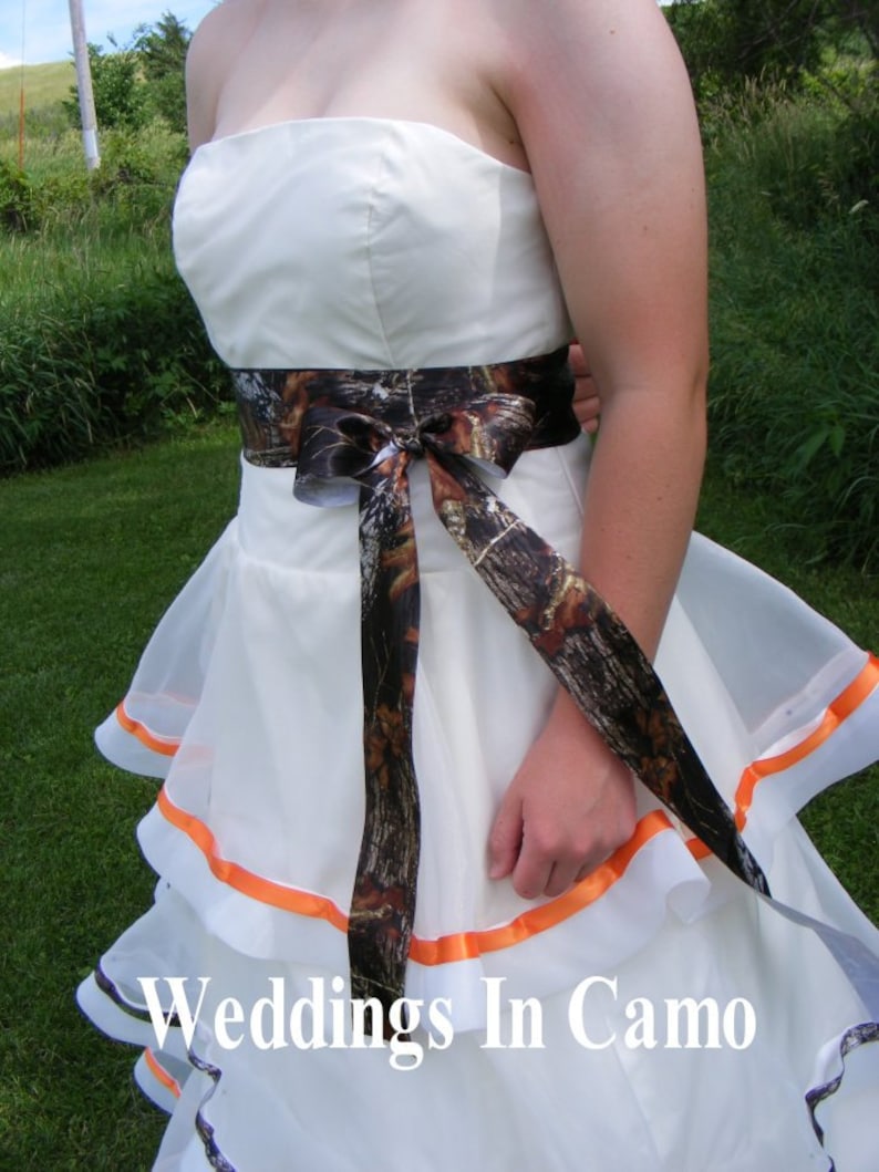 May include: A white wedding dress with a camouflage ribbon tied around the waist. The dress has a layered skirt with orange trim.