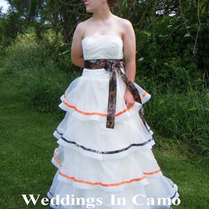 May include: A white wedding dress with a camouflage and orange ribbon sash. The dress has multiple layers of tulle and is photographed outdoors in a grassy area.