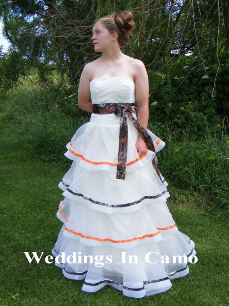 May include: A white wedding dress with a camouflage and orange ribbon sash. The dress has multiple layers of tulle and is photographed outdoors in a grassy area.