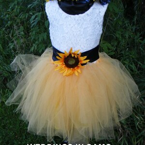 May include: A white lace sleeveless dress with a black satin sash and a sunflower accent. The dress has a yellow tulle skirt and a sunflower and blue flower crown.