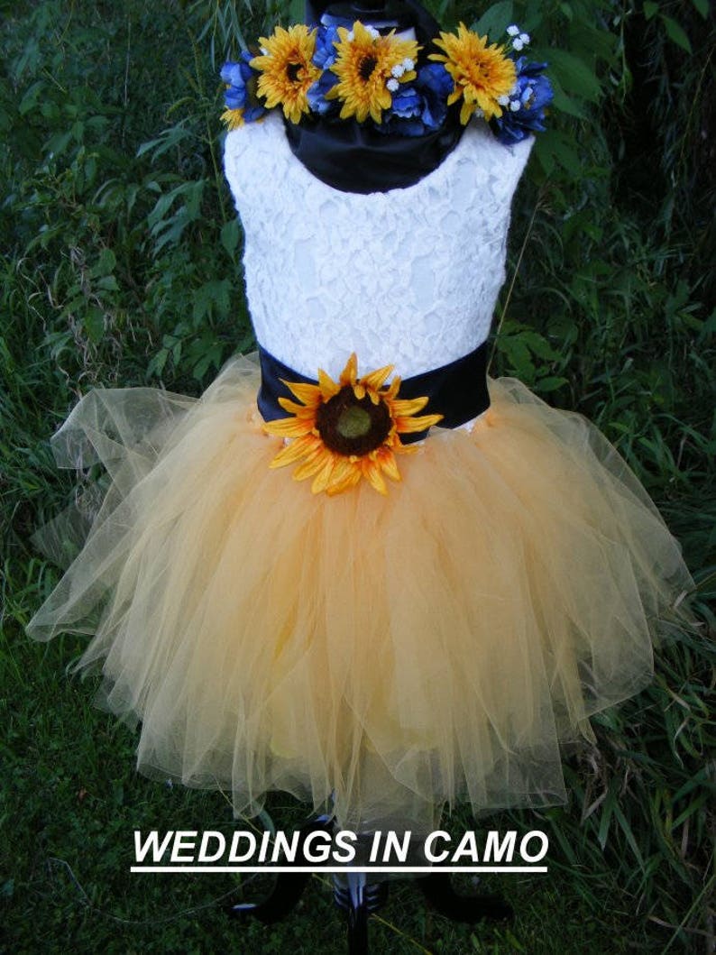 May include: A white lace sleeveless dress with a black satin sash and a sunflower accent. The dress has a yellow tulle skirt and a sunflower and blue flower crown.