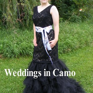 May include: A black lace wedding dress with a white camouflage ribbon belt and a black tulle skirt. The dress is worn by a person standing in a grassy area. The text "Weddings in Camo" is visible at the bottom of the image.