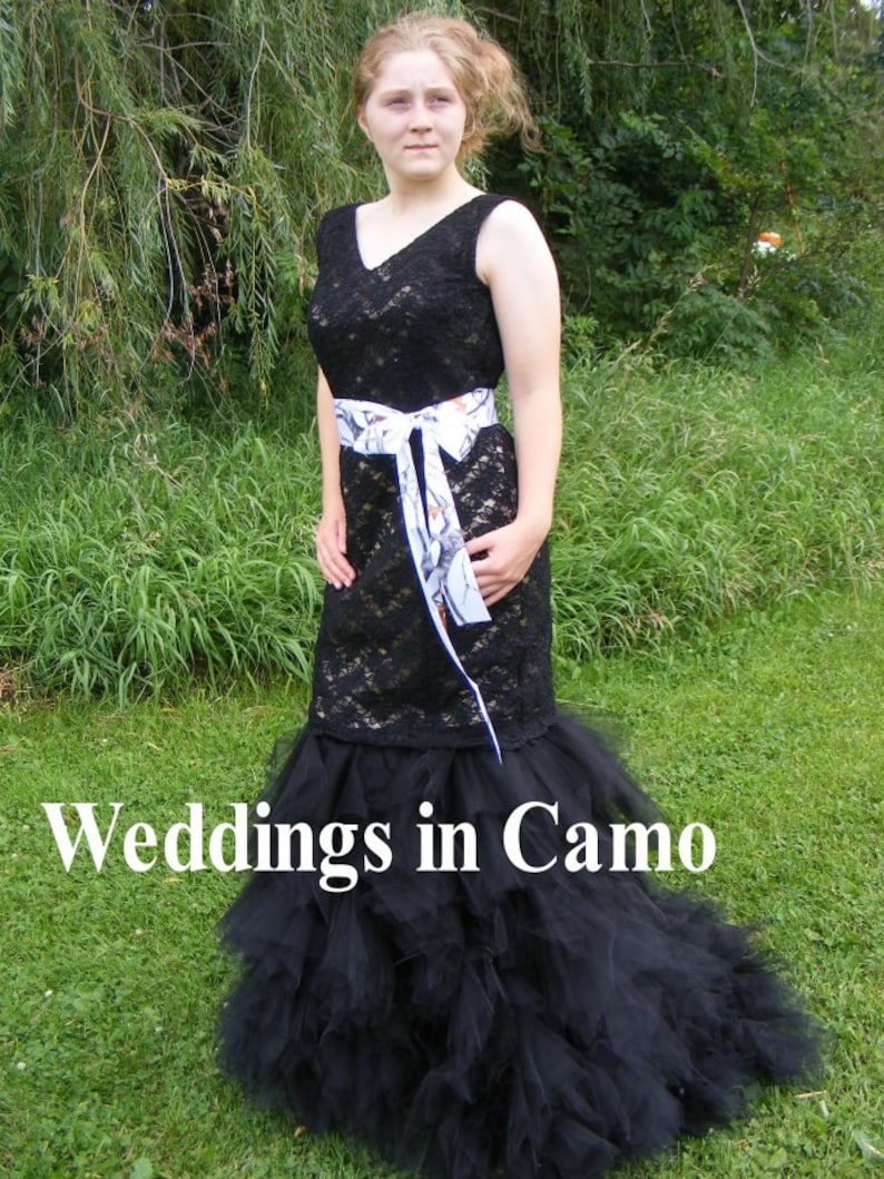 May include: A black lace wedding dress with a white camouflage ribbon belt and a black tulle skirt. The dress is worn by a person standing in a grassy area. The text "Weddings in Camo" is visible at the bottom of the image.