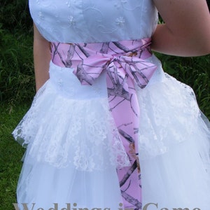 May include: A white strapless wedding dress with a pink and camouflage ribbon tied around the waist. The dress has a lace overlay and a tulle skirt.