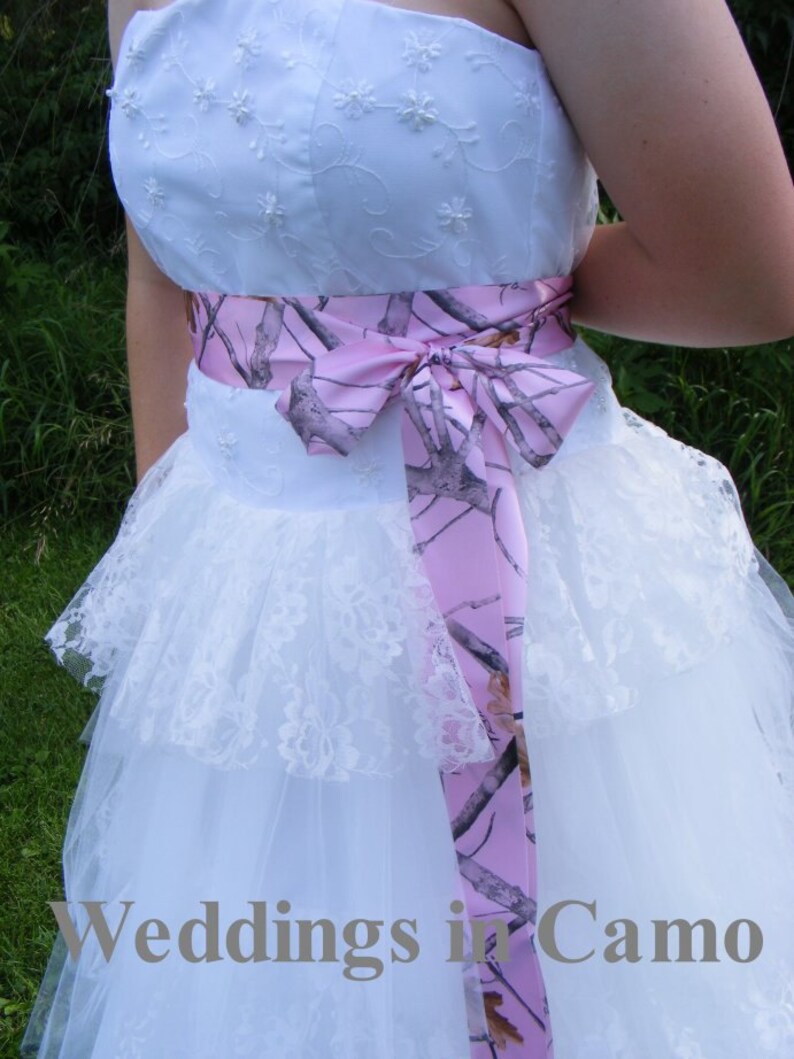 May include: A white strapless wedding dress with a pink and camouflage ribbon tied around the waist. The dress has a lace overlay and a tulle skirt.