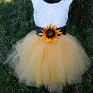 May include: A white lace sleeveless dress with a black satin sash and a sunflower accent. The dress has a yellow tulle skirt. The dress is on a mannequin and the background is green.