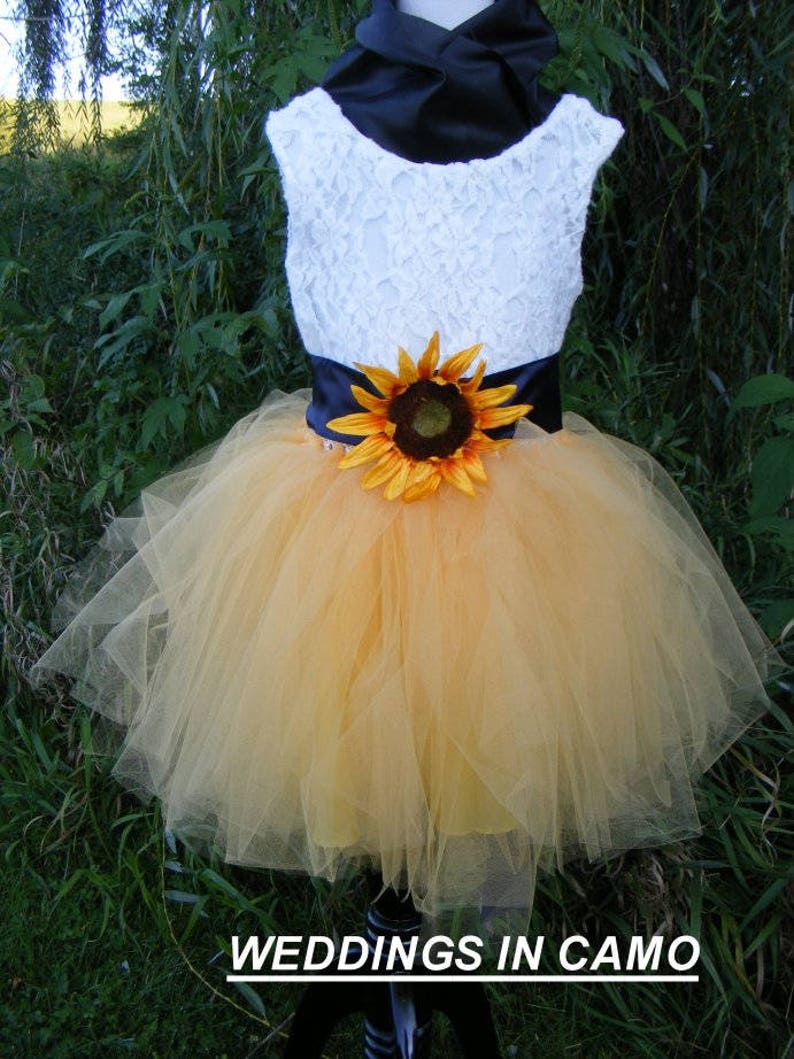 May include: A white lace sleeveless dress with a black satin sash and a sunflower accent. The dress has a yellow tulle skirt. The dress is on a mannequin and the background is green.