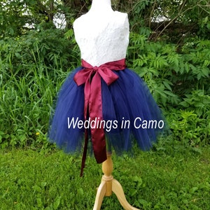 May include: A navy blue tulle skirt with a white lace bodice and a burgundy satin sash. The skirt is on a mannequin and the text "Weddings in Camo" is visible on the skirt.