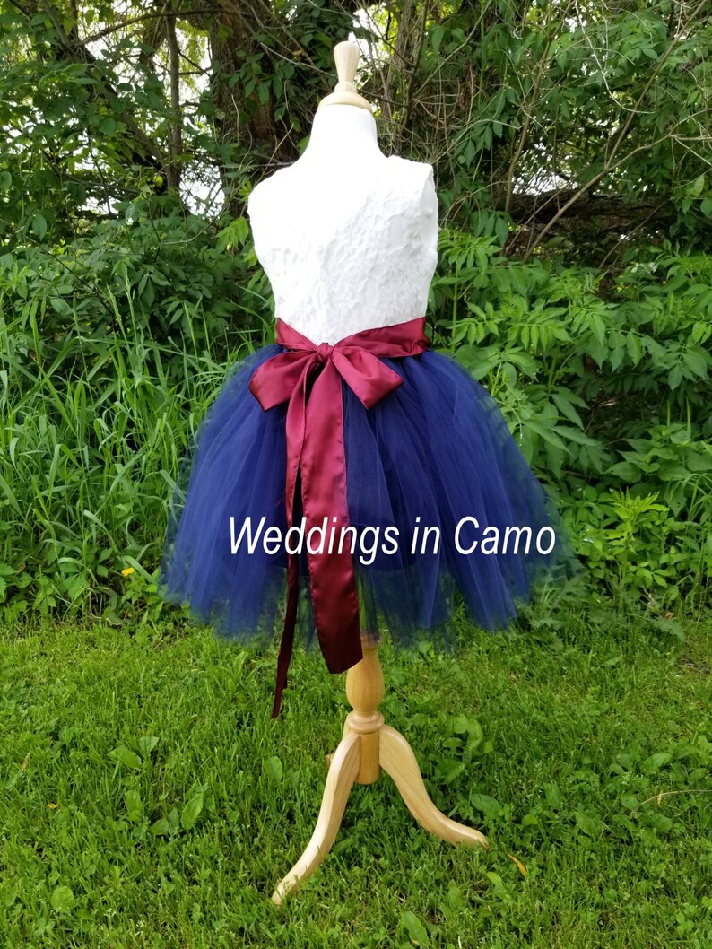 May include: A navy blue tulle skirt with a white lace bodice and a burgundy satin sash. The skirt is on a mannequin and the text "Weddings in Camo" is visible on the skirt.