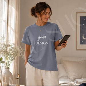 May include: A slate blue short-sleeved t-shirt featuring the white text "your DESIGN" printed on the chest. The shirt has a crew neckline and a relaxed fit, paired with light beige trousers. The garment is shown in a neutral indoor setting.