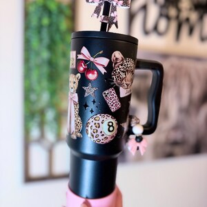 May include: Black tumbler with a pink base and handle, decorated with leopard print and cherry decals. A bow-shaped topper and a charm with pink accents add flair. The tumbler features a black straw.