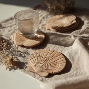 Peut inclure: Sous-verres en bois en forme de coquillages. Un verre transparent rempli d'eau repose sur un sous-verre. Les sous-verres sont de couleur marron clair et présentent un motif de coquillage sculpté. Une pile de sous-verres est à l'arrière-plan.