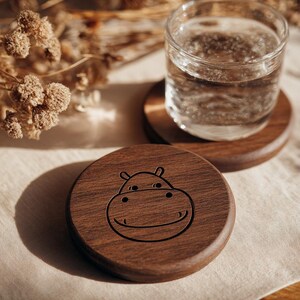May include: Wooden coasters with a hippo face design. One coaster has a glass of water on it. The coasters are made of dark brown wood and are round. The hippo face is etched into the wood. The background is a light beige fabric.