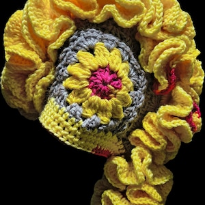 May include: A handmade crochet hat featuring a central flower motif with a bright pink center, yellow petals, and gray surrounding stitches. The hat is adorned with a large, voluminous, ruffled yellow border that cascades down one side.