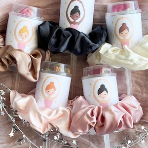 Op de afbeelding: Verschillende witte containers met doorzichtige deksels, elk met een ballerina-illustratie, worden omringd door satijnen scrunchies in verschillende kleuren. De containers zijn gevuld met kleine items. Er is ook een zilveren sterrenslingers aanwezig.