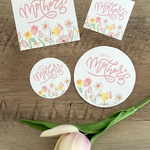 May include: A collection of Mother's Day cards and coasters with floral designs. The cards and coasters feature the text "Happy Mother's Day" in pink script, with a border of watercolor flowers in yellow, pink, and white.