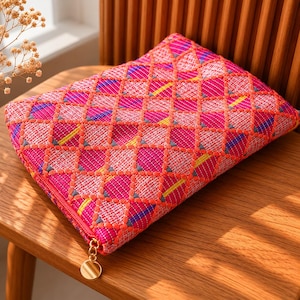 May include: A rectangular zippered pouch featuring a vibrant pink, orange, and white woven diamond pattern. The fabric has a textured finish with small embroidered details in yellow and blue. A gold-toned circular metal zipper pull is attached.