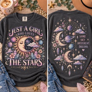 May include: A charcoal gray t-shirt featuring a celestial graphic. The front displays a sun and moon face with planets, flowers, and the text "JUST A GIRL WHO LOVES THE STARS." The back shows a moon, planets, crystals, and "SAME SKY DIFFERENT DREAMS."