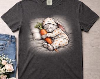 Puede incluir: Camiseta gris oscuro con una ilustración de un conejito durmiendo en pijama, abrazando una zanahoria. El conejito está sobre una almohada. La camiseta tiene una etiqueta Comfort Colors. El diseño es detallado e ideal para el uso diario.