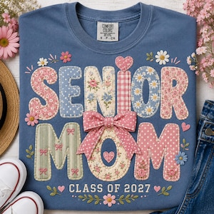 May include: A blue t-shirt featuring the text SENIOR MOM in large, fabric-appliqued letters with various floral, polka dot, and gingham patterns. A pink fabric bow is centered over the letters, with the text CLASS OF 2027 printed below.