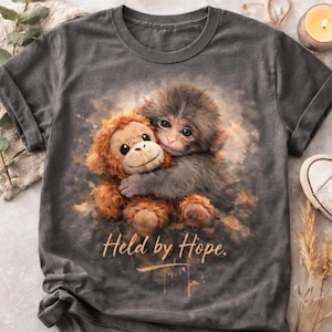 May include: Dark gray t-shirt featuring a detailed illustration of a baby monkey hugging a stuffed animal monkey. The text "Held by Hope" is printed in a cursive font below the image. The shirt has short sleeves and a crew neck.