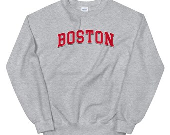 Op de afbeelding: Een grijze sweatshirt met het woord "BOSTON" in rood, omlijnd met wit, over de borst. De sweatshirt heeft een ronde hals en lange mouwen, geschikt voor vrijetijdskleding. Het Gildan-merklabel is zichtbaar.