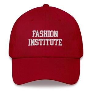 May include: A red baseball cap with the words "FASHION INSTITUTE" embroidered in white across the front. The cap has a curved brim and a rounded crown, suitable for casual wear.