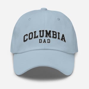 May include: A light blue baseball cap with the word "COLUMBIA" arched across the front in black, above the word "DAD". The cap has a curved brim and an adjustable strap.
