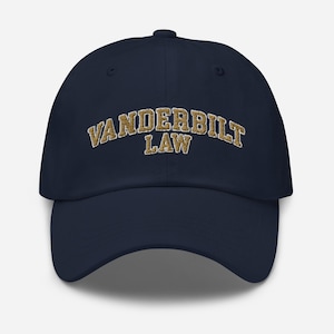 May include: A navy blue baseball cap with the text "VANDERBILT LAW" embroidered in gold-colored thread. The cap has a curved brim and a structured crown, suitable for everyday wear.