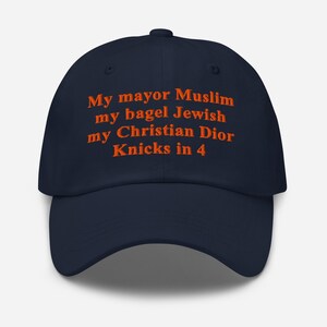 May include: A navy blue baseball cap featuring orange embroidered text that reads: My mayor Muslim, my bagel Jewish, my Christian Dior, Knicks in 4. The cap has a structured crown, a curved brim, and a button at the top center of the fabric panels.