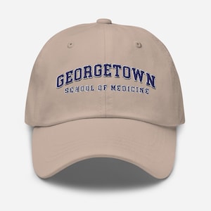 May include: A tan baseball cap with the text "GEORGETOWN SCHOOL OF MEDICINE" embroidered in navy blue. The cap has a curved brim and a button on top.