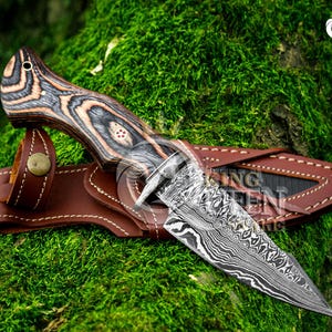 May include: A Damascus steel knife with a wooden handle and a brown leather sheath. The handle features a wood grain pattern in shades of gray, brown, and black. The knife is resting on a bed of green moss.