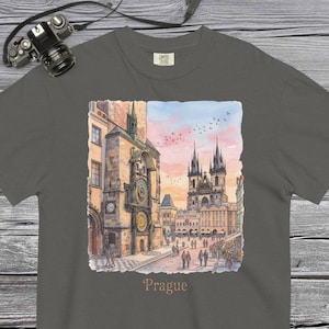 Pode incluir: T-shirt cinzenta escura com uma ilustração em aquarela do horizonte de Praga, incluindo o Relógio Astronómico e a Igreja de Nossa Senhora de Týn. A palavra "Prague" está impressa abaixo da imagem. Uma câmera está no canto superior esquerdo.