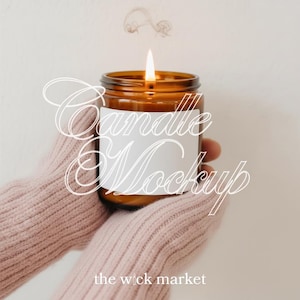 May include: A lit amber glass candle jar with a white label is held by someone wearing a pink sweater. The candle flame is burning brightly, with wisps of smoke rising. The text "Candle Mockup" is overlaid on the image, along with "the wick market".