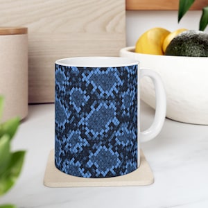 May include: White ceramic mug featuring a blue snakeskin pattern. The mug has a white handle and is sitting on a beige coaster. The mug is filled with a dark liquid. The mug is a kitchen and dining accessory.