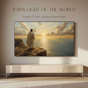 May include: A digital television display showing a landscape painting titled JESUS LIGHT OF THE WORLD. The artwork depicts a figure in a robe standing on a rocky cliff overlooking a vast ocean at sunrise with golden light rays and textured clouds.