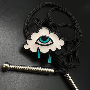 May include: A black bolo tie featuring a white cloud pendant with a teal eye design and teal teardrops. The bolo tie has a black braided cord and a silver-toned metal slide and tips. The pendant is approximately 1.5 inches wide.