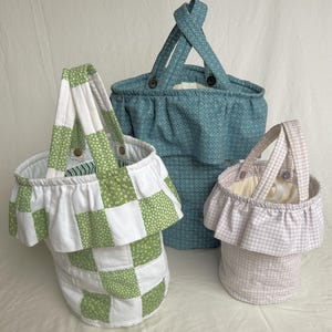 May include: Three fabric baskets in different sizes. The largest is teal with a geometric pattern and button fastenings. The medium basket is green and white patchwork. The smallest is pink gingham with a ruffled edge and a handle.