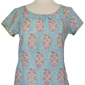 Pode incluir: Uma blusa azul turquesa de manga curta com estampa paisley em tons de marrom, vermelho e branco. A blusa tem uma borda verde ao redor do decote e das mangas.