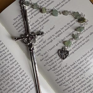 Puede incluir: Un marcap&aacute;ginas plateado con dise&ntilde;o de espada y una cadena de cuentas con piedras verdes descansa sobre un libro abierto. Un dije en forma de coraz&oacute;n con una cruz cuelga de la cadena. El libro est&aacute; abierto en una p&aacute;gina de texto.